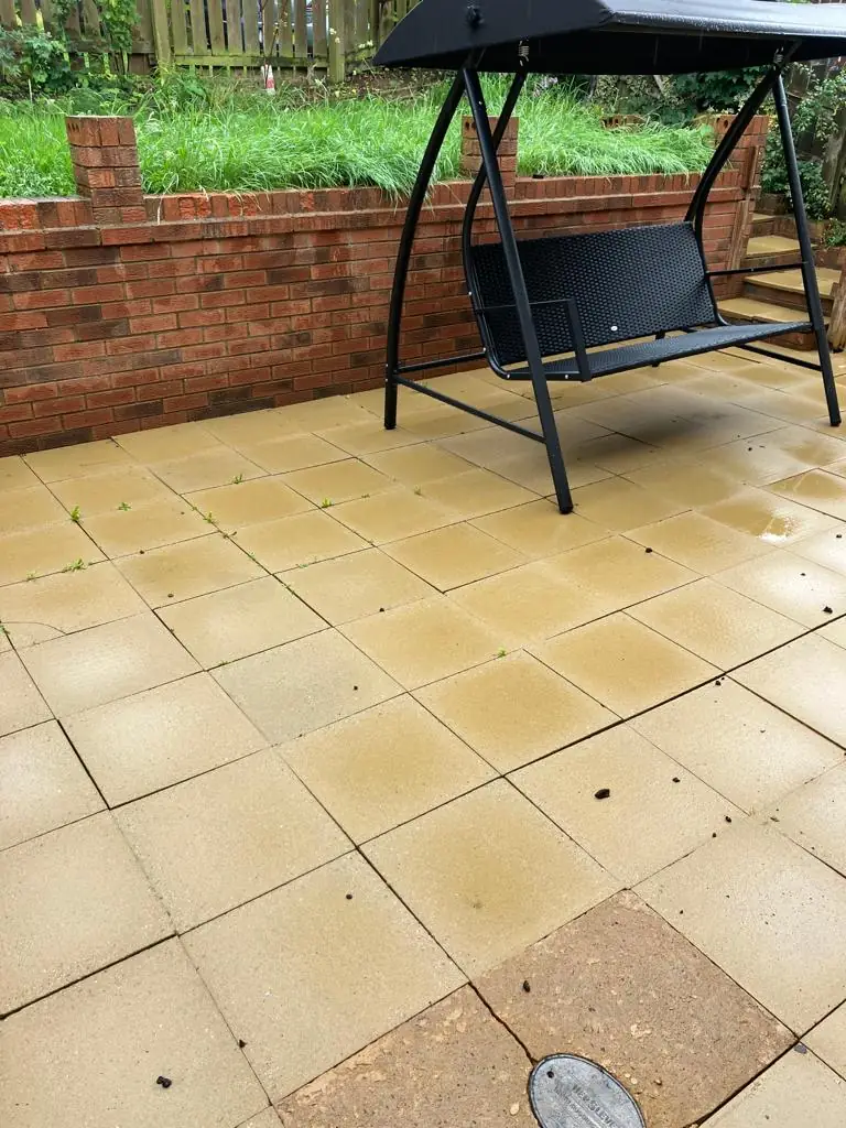 Clean buff-yellow patio paving beside a red brick wall with a black garden bench after professional cleaning