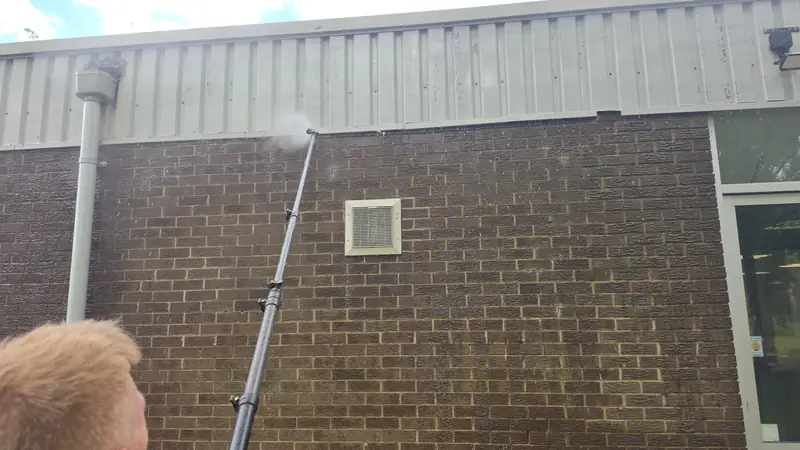Operator from behind directing a soft wash jet onto a wall-mounted ventilation grille, sun catching blond hair