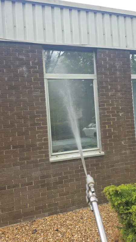 Soft wash spray reaching a tall window and surrounding brickwork, a car visible reflected in the glass