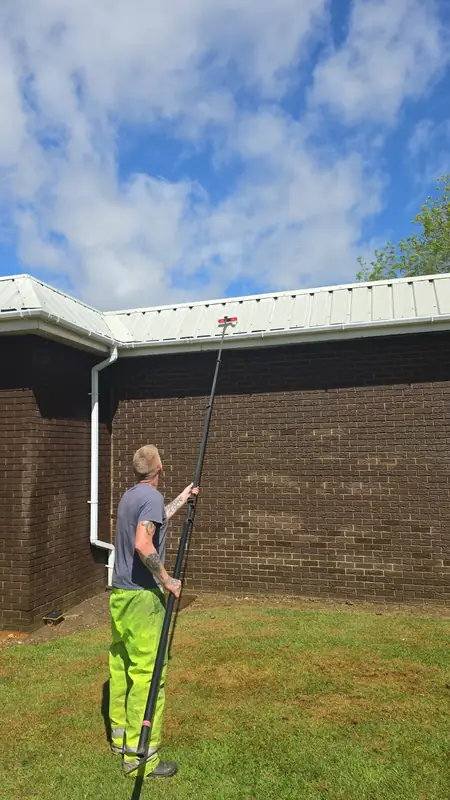 Operator in hi-vis using a water-fed telescopic pole to clean the gable cladding while standing on the lawn