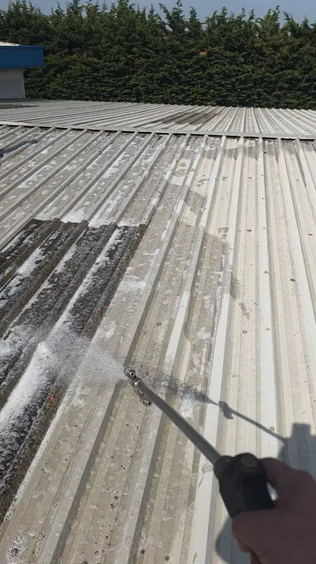 Operator hand holding a low-pressure spray wand applying vertical streaks of biocide foam down a corrugated metal roof panel with the blue parapet behind