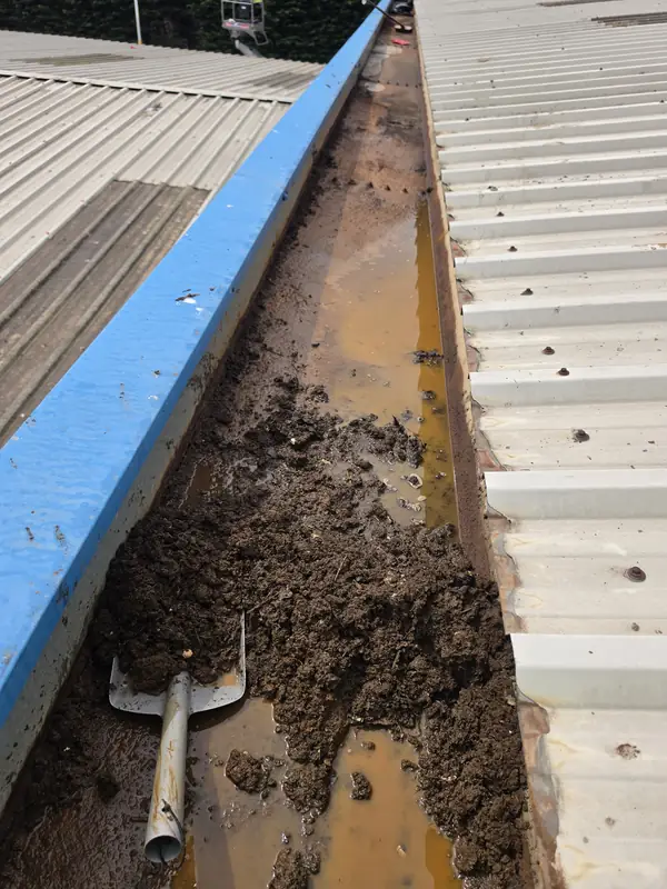 Close view of a gutter completely filled with brown mud and organic debris with a red and white plastic scoop blade resting in the middle