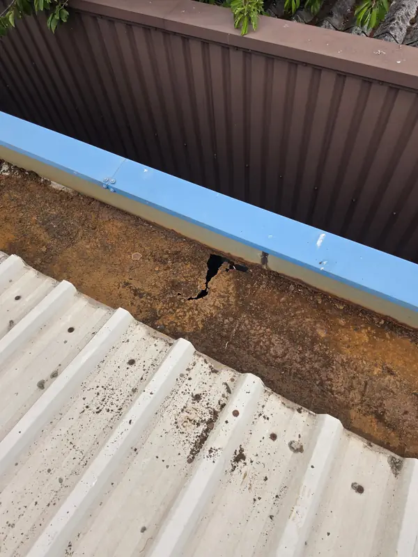 Dramatic close-up of an irregular star-shaped hole rusted through the gutter metal with brown timber fascia visible below