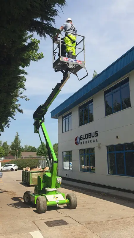 Nifty articulated boom MEWP extended with the operator in a white helmet on the basket and the Globus Medical UK logo and blue-trim windows clearly visible on the building behind