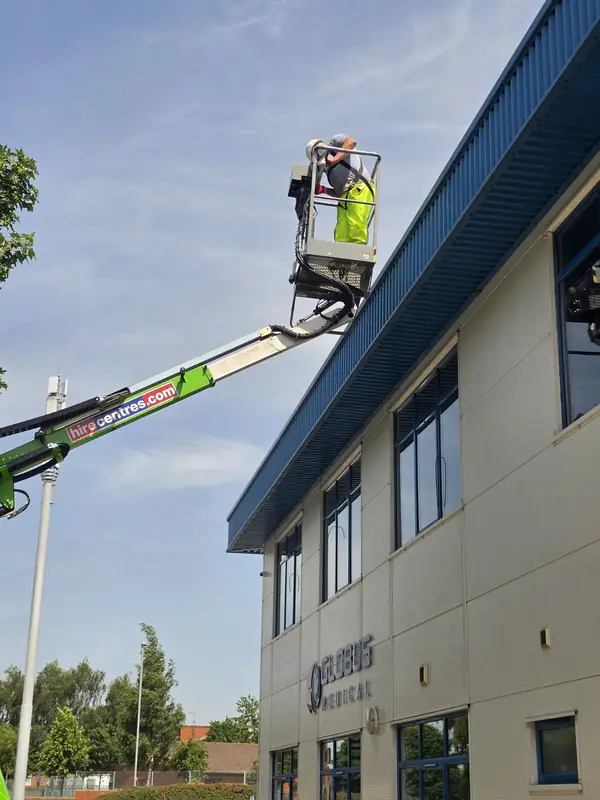 Low-angle view looking up at the operator on the MEWP basket with the green hire-centre boom against sky and the Globus Medical facade behind