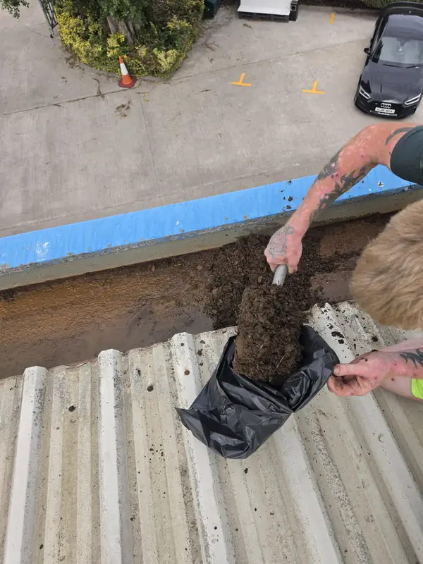 First-person view of both gloved hands holding a white plastic scoop loaded with mud above an open black debris bag on the roof panel