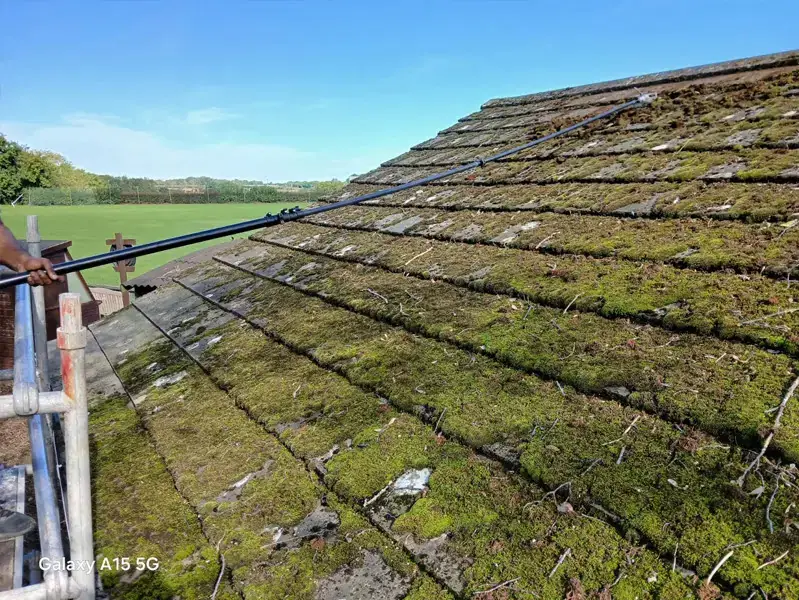 Operative scraping heavy green moss from a roof viewed from scaffold level with open countryside behind