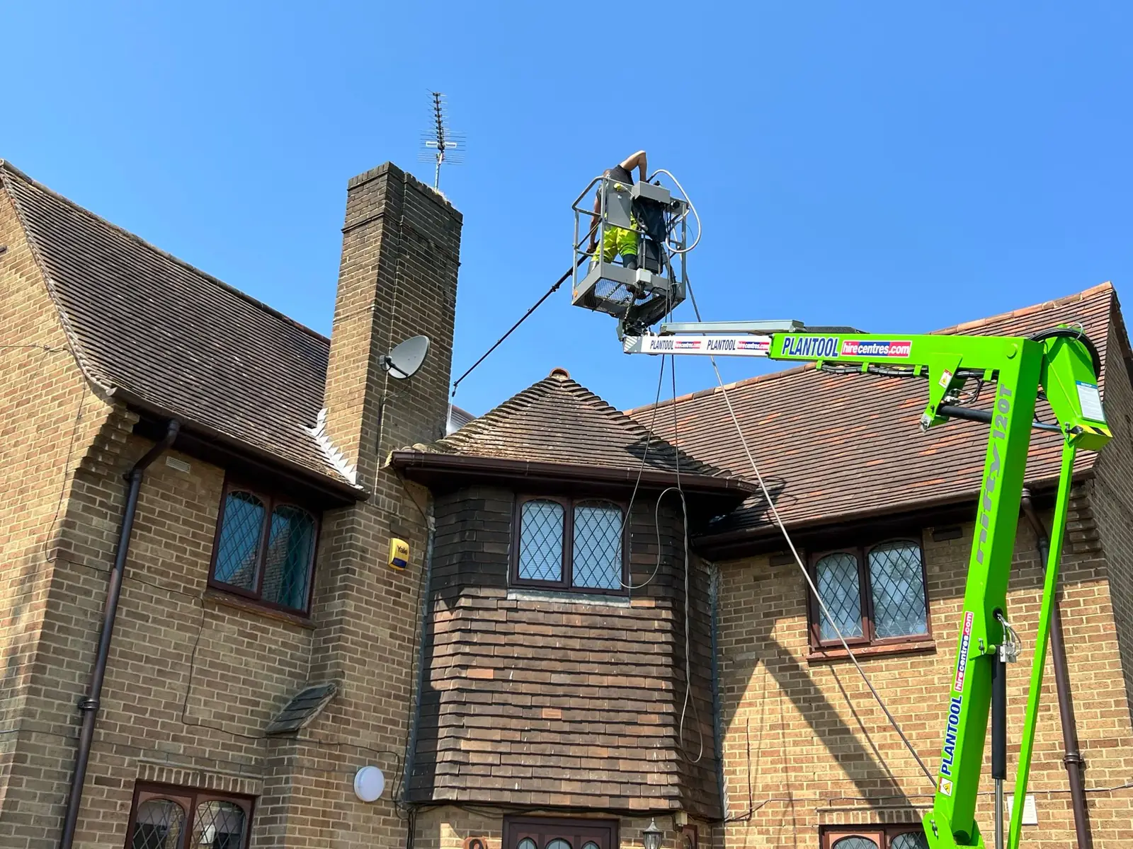 Operative cleaning the brown roof of a detached brick house from a green PLANTOOL cherry picker against a blue sky