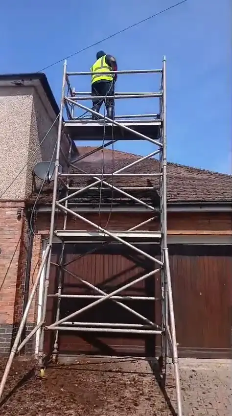 Hi-vis operative scraping moss from a roof on a scaffold tower beside a brick house and timber garage