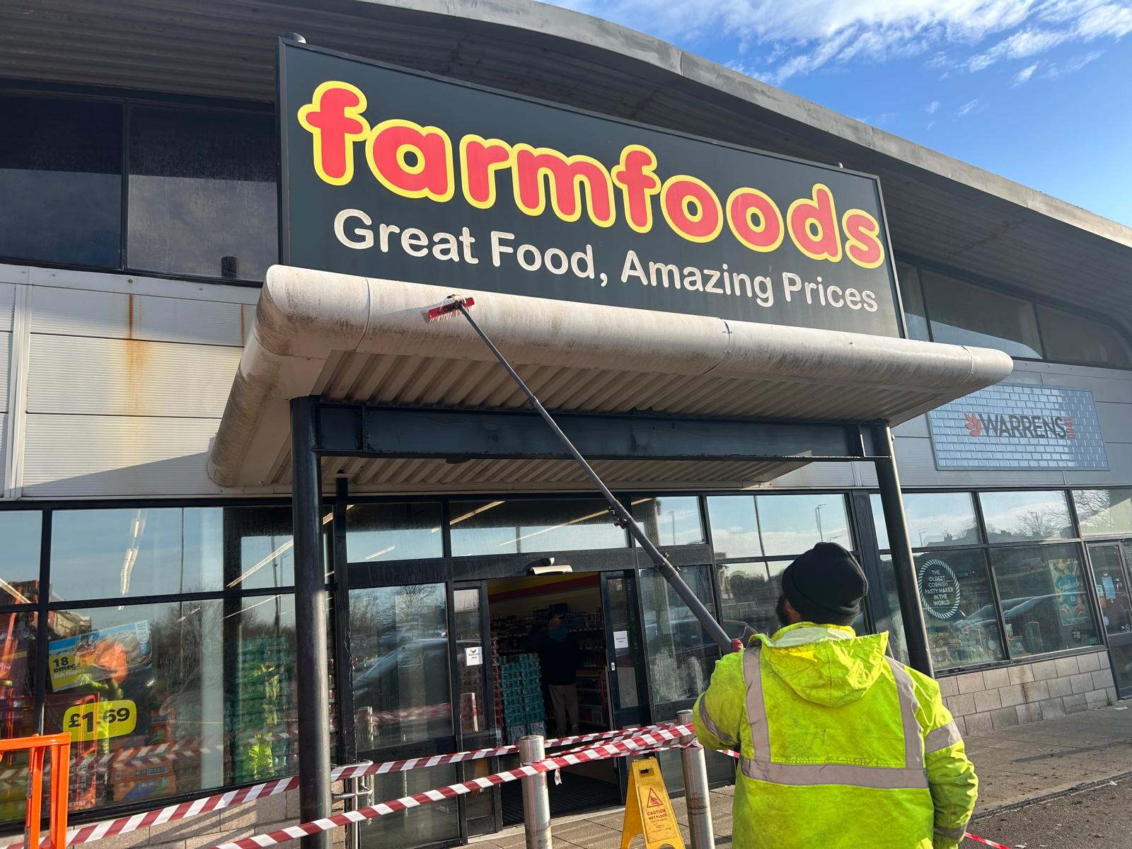 Professional cleaner using telescopic lance to pressure wash commercial supermarket entrance canopy showing cleaning in progress at Farmfoods